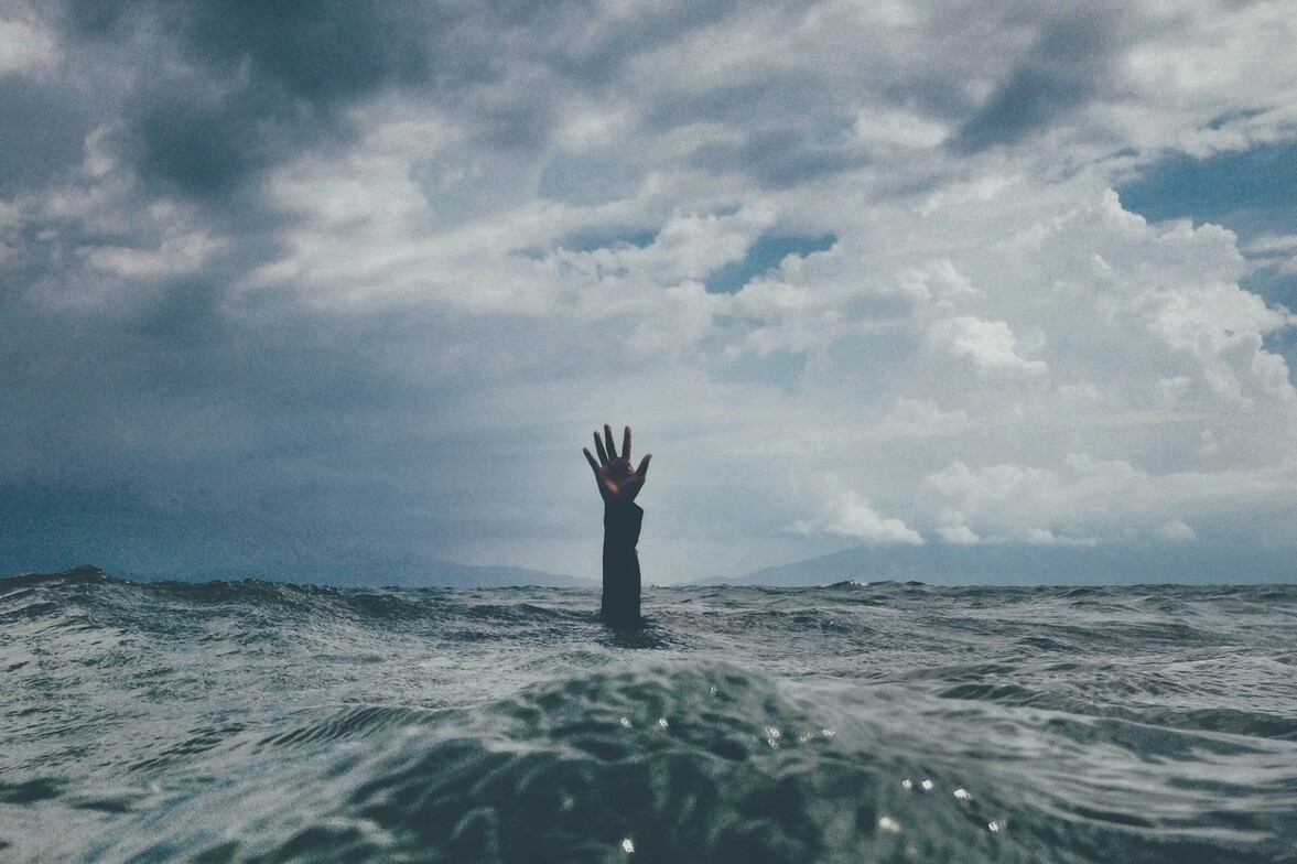 A single hand reaches up from beneath the surface of a body of water, symbolizing the feeling of drowning in stress and overwhelm